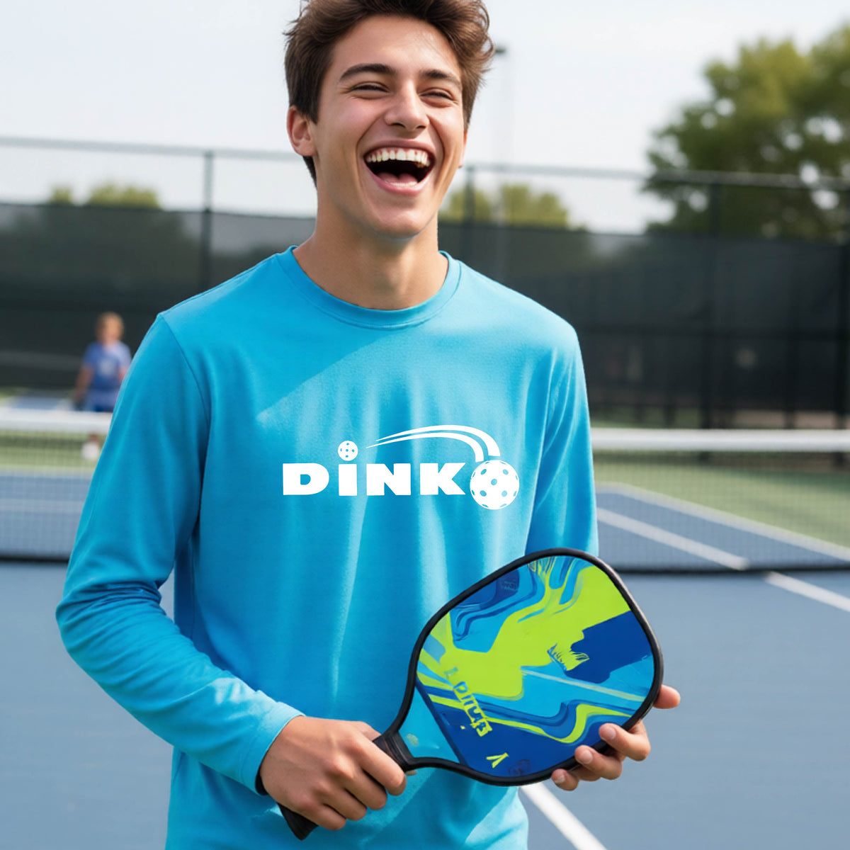 A young person wearing a light blue long-sleeve t-shirt with a pickleball design of text 'DINK' with a white pickleall in flight, standing on a pickleball court
