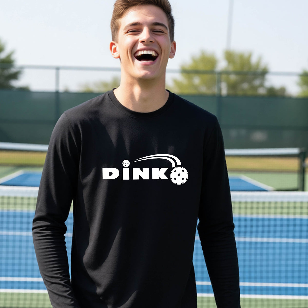 A young person wearing a black long-sleeve t-shirt with a pickleball design of text 'DINK' with a white pickleall in flight, standing on a pickleball court