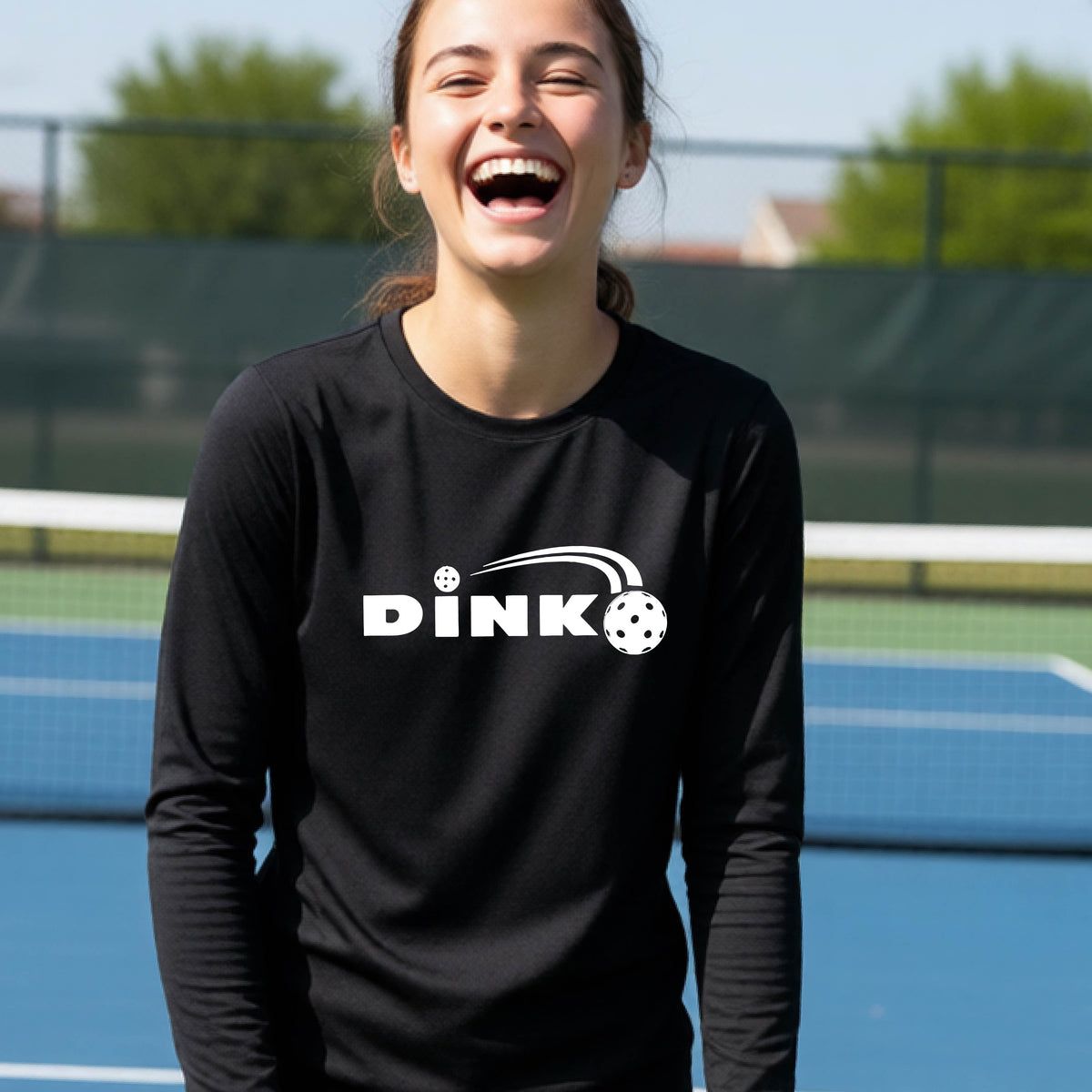A young person wearing a black long-sleeve t-shirt with a pickleball design of text 'DINK' with a white pickleall in flight, standing on a pickleball court
