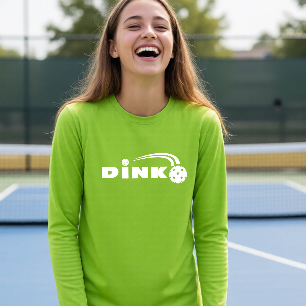 A young person wearing a lime green long-sleeve t-shirt with a pickleball design of text 'DINK' with a white pickleall in flight, standing on a pickleball court