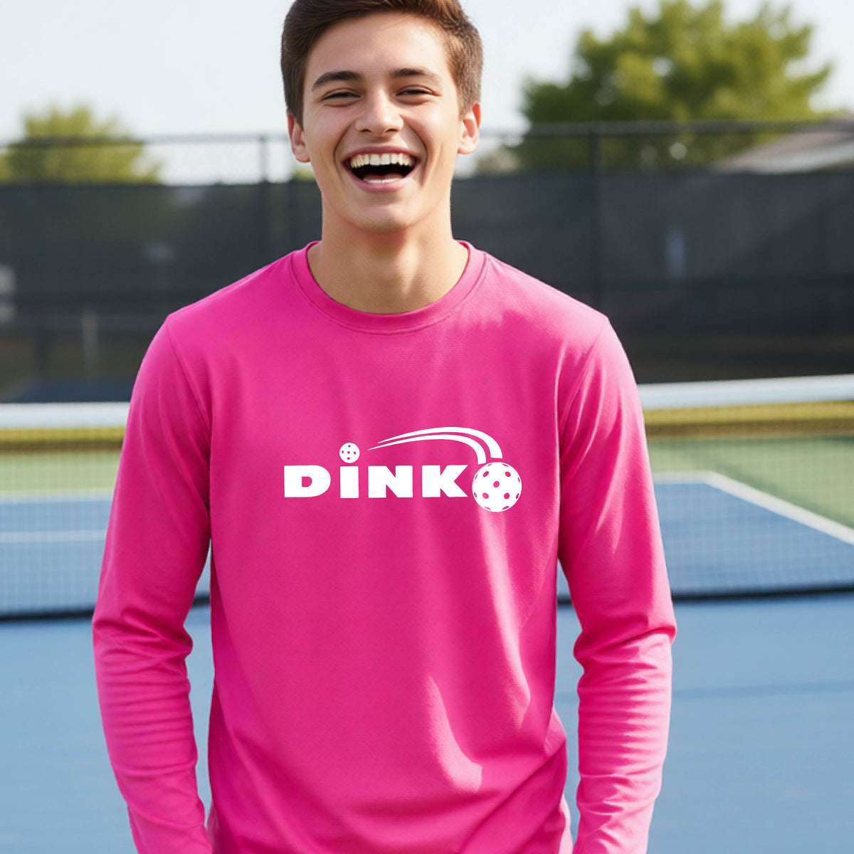 A young person wearing a pink long-sleeve t-shirt with a pickleball design of text 'DINK' with a white pickleall in flight, standing on a pickleball court