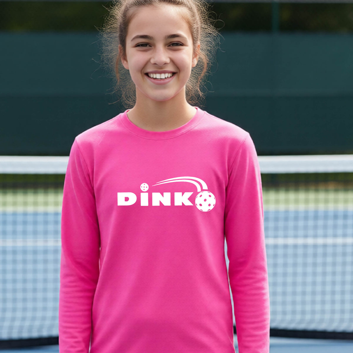 A young person wearing a pink long-sleeve t-shirt with a pickleball design of text 'DINK' with a white pickleall in flight, standing on a pickleball court.