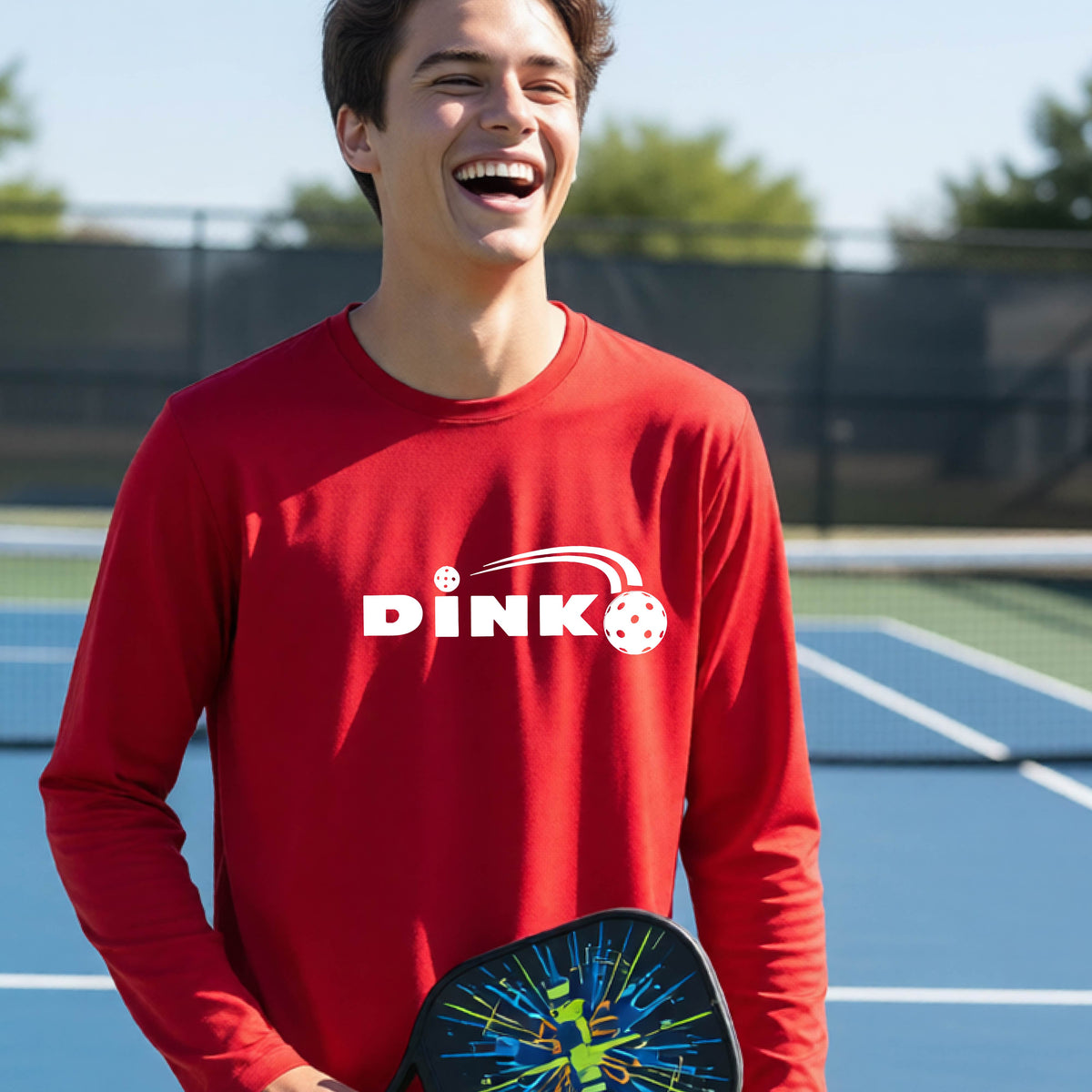 A young person wearing a red long-sleeve t-shirt with a pickleball design of text 'DINK' with a white pickleall in flight, standing on a pickleball court