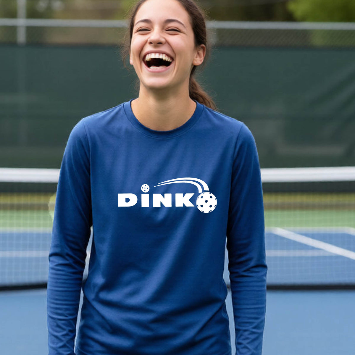 A young person wearing a royal blue long-sleeve t-shirt with a pickleball design of text 'DINK' with a white pickleall in flight, standing on a pickleball court
