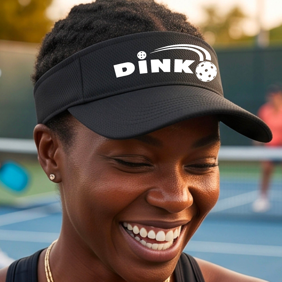 A close-up of a black visor with a graphic of white 'DINK' and a dink pickleball logo printed in white on the front, set against a blurred outdoor pickleball court background.