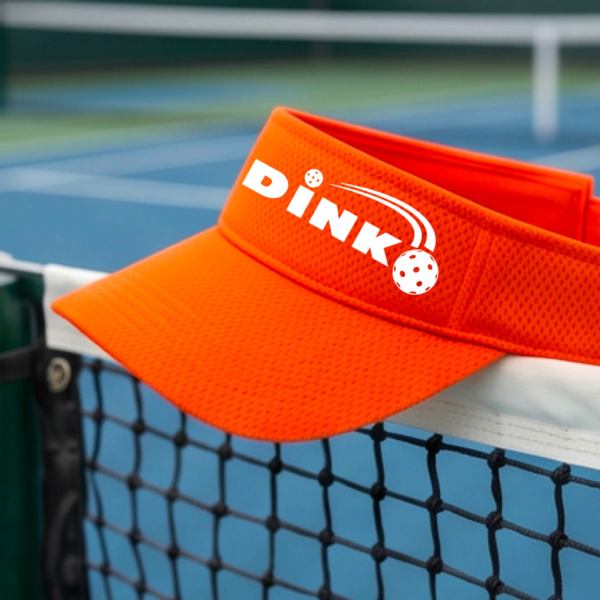 A close-up of an orange visor with a graphic of white 'DINK' and a dink pickleball logo printed in white on the front, set against a blurred outdoor pickleball court background.