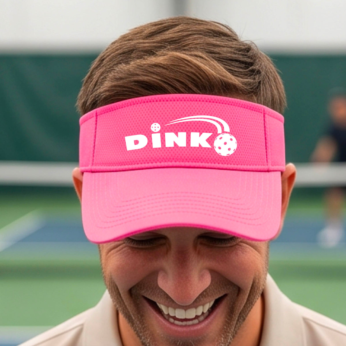 A close-up of a pink visor with a graphic of white 'DINK' and a dink pickleball logo printed in white on the front, set against a blurred outdoor pickleball court background.