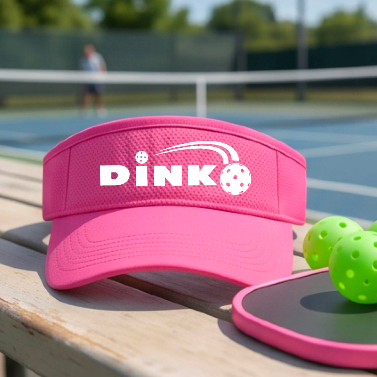 A close-up of a pink visor with a graphic of white 'DINK' and a dink pickleball logo printed in white on the front, set against a blurred outdoor pickleball court background.