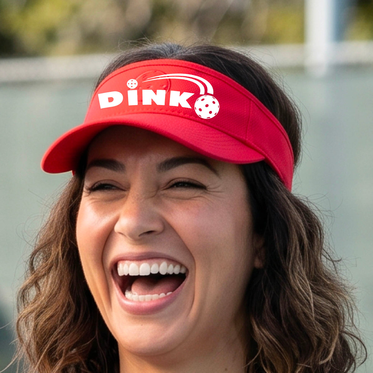 A close-up of a red visor with a graphic of white 'DINK' and a dink pickleball logo printed in white on the front, set against a blurred outdoor pickleball court background.