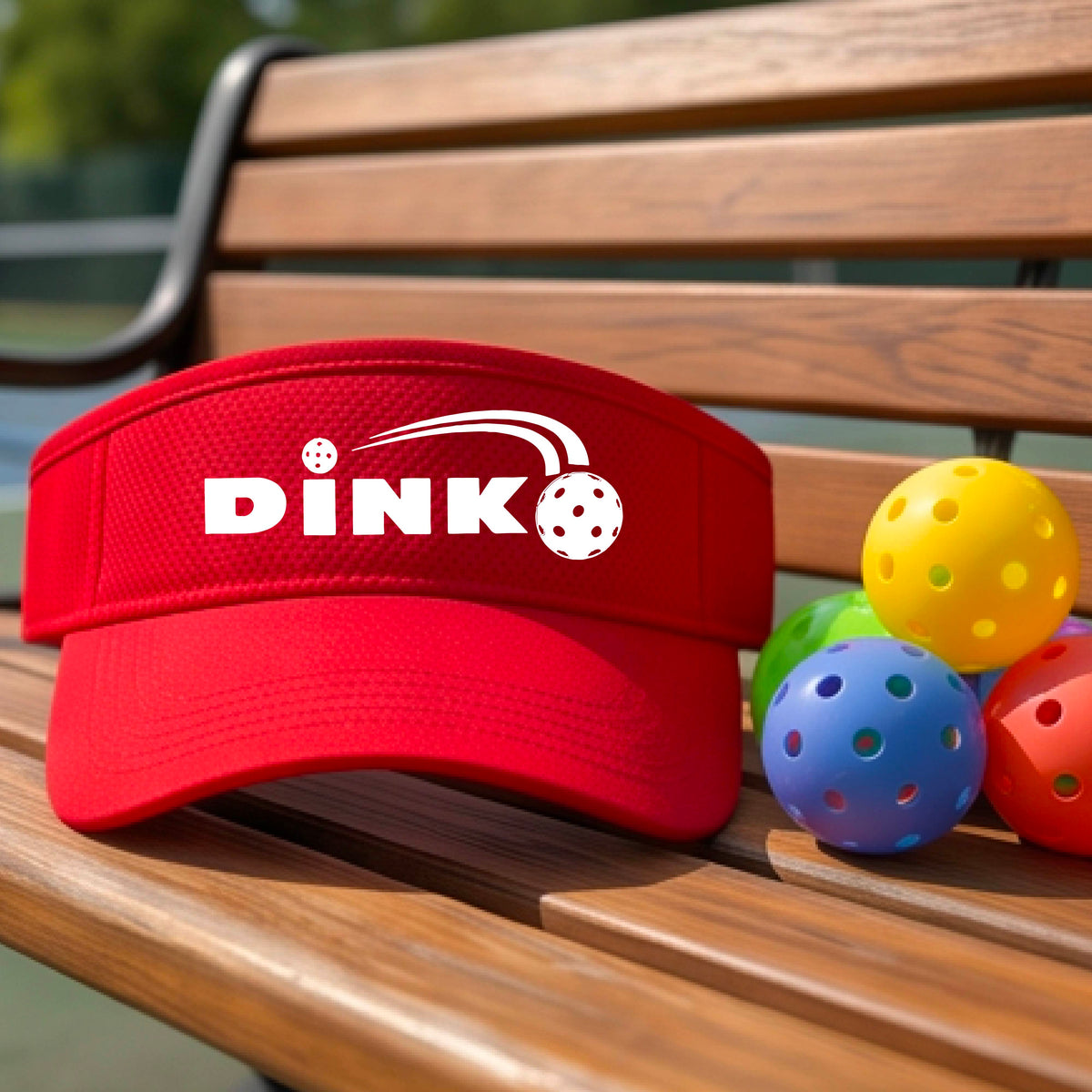 A close-up of a red visor with a graphic of white 'DINK' and a dink pickleball logo printed in white on the front, set against a blurred outdoor pickleball court background.