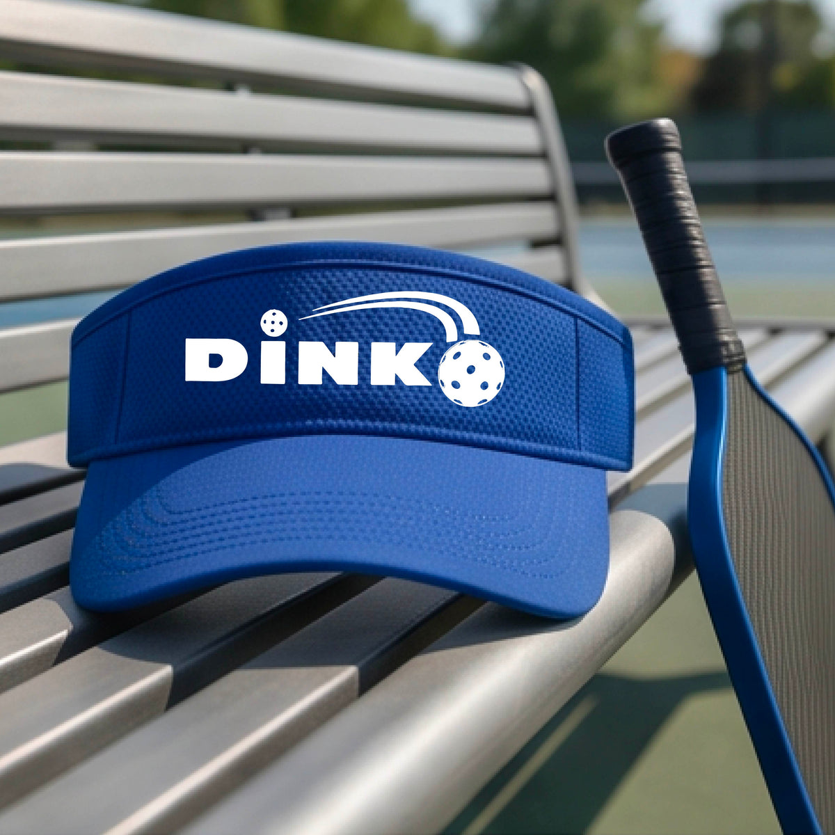 A close-up of a royal blue visor with a graphic of white 'DINK' and a dink pickleball logo printed in white on the front, set against a blurred outdoor pickleball court background.