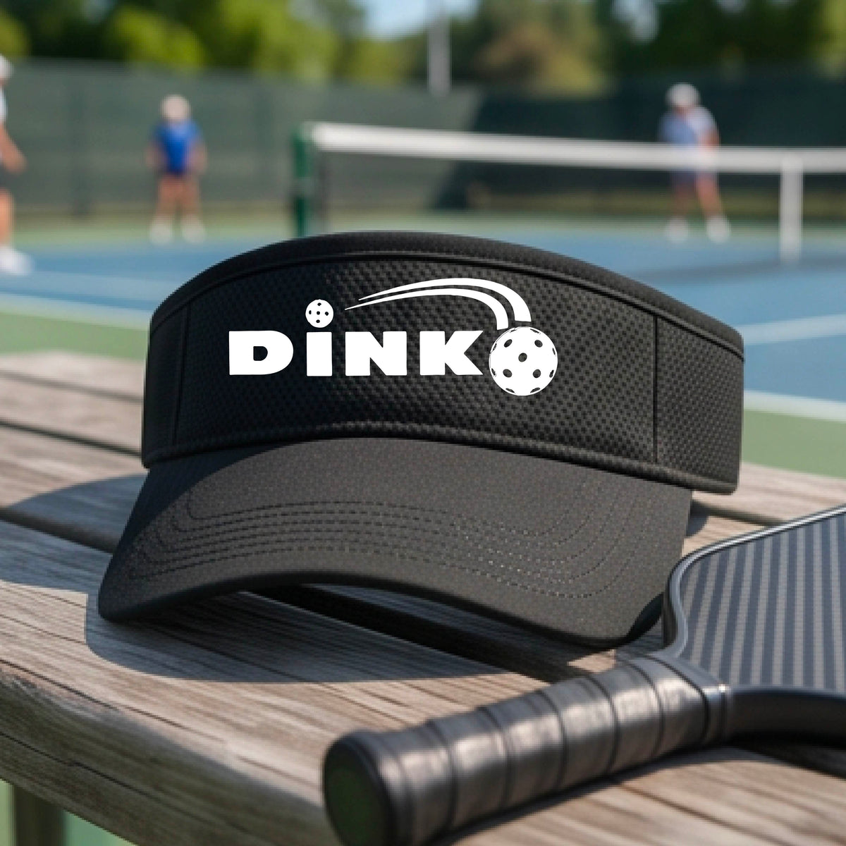 A close-up of a black visor with a graphic of white 'DINK' and a dink pickleball logo printed in white on the front, set against a blurred outdoor pickleball court background.