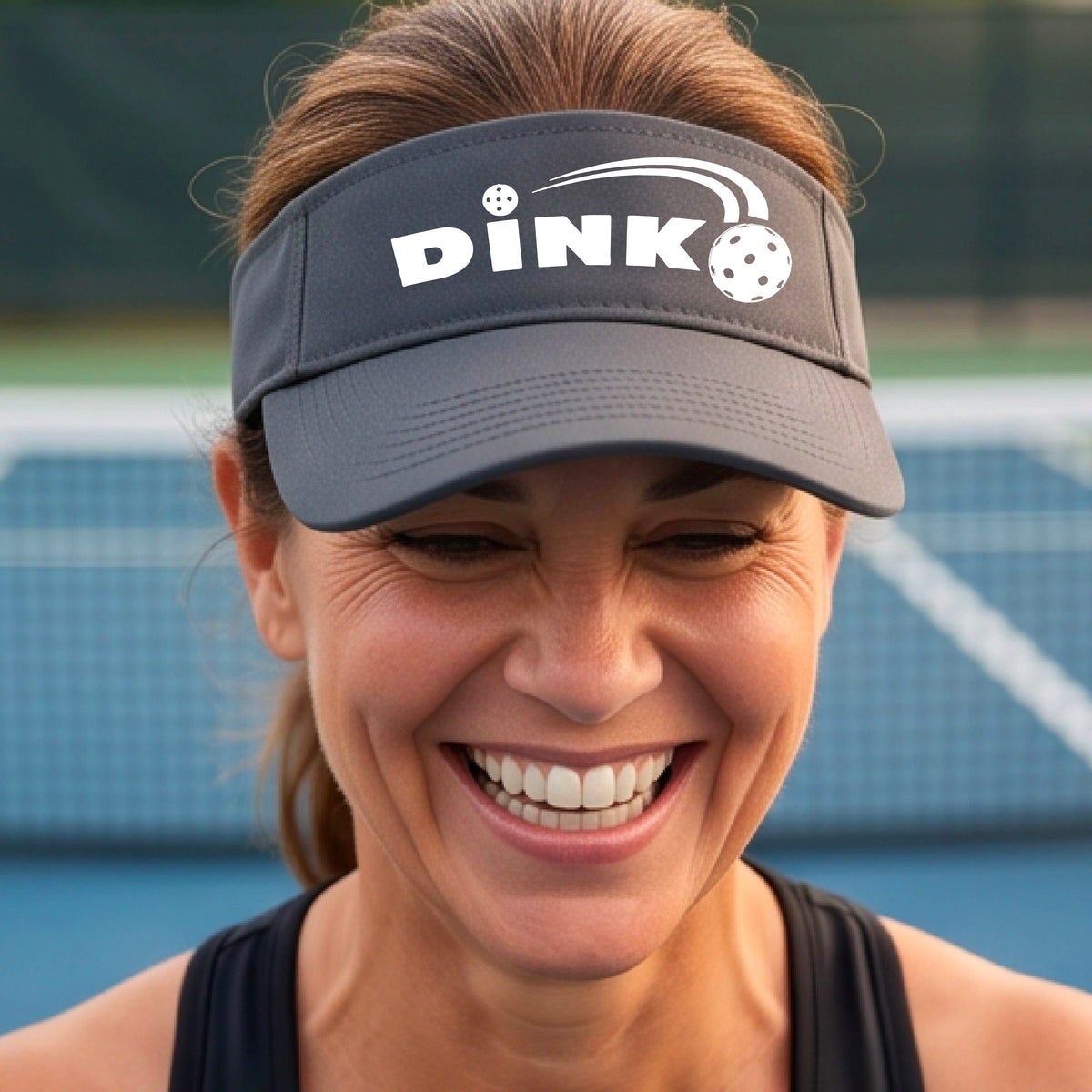 A close-up of a gray visor with a graphic of white 'DINK' and a dink pickleball logo printed in white on the front, set against a blurred outdoor pickleball court background.