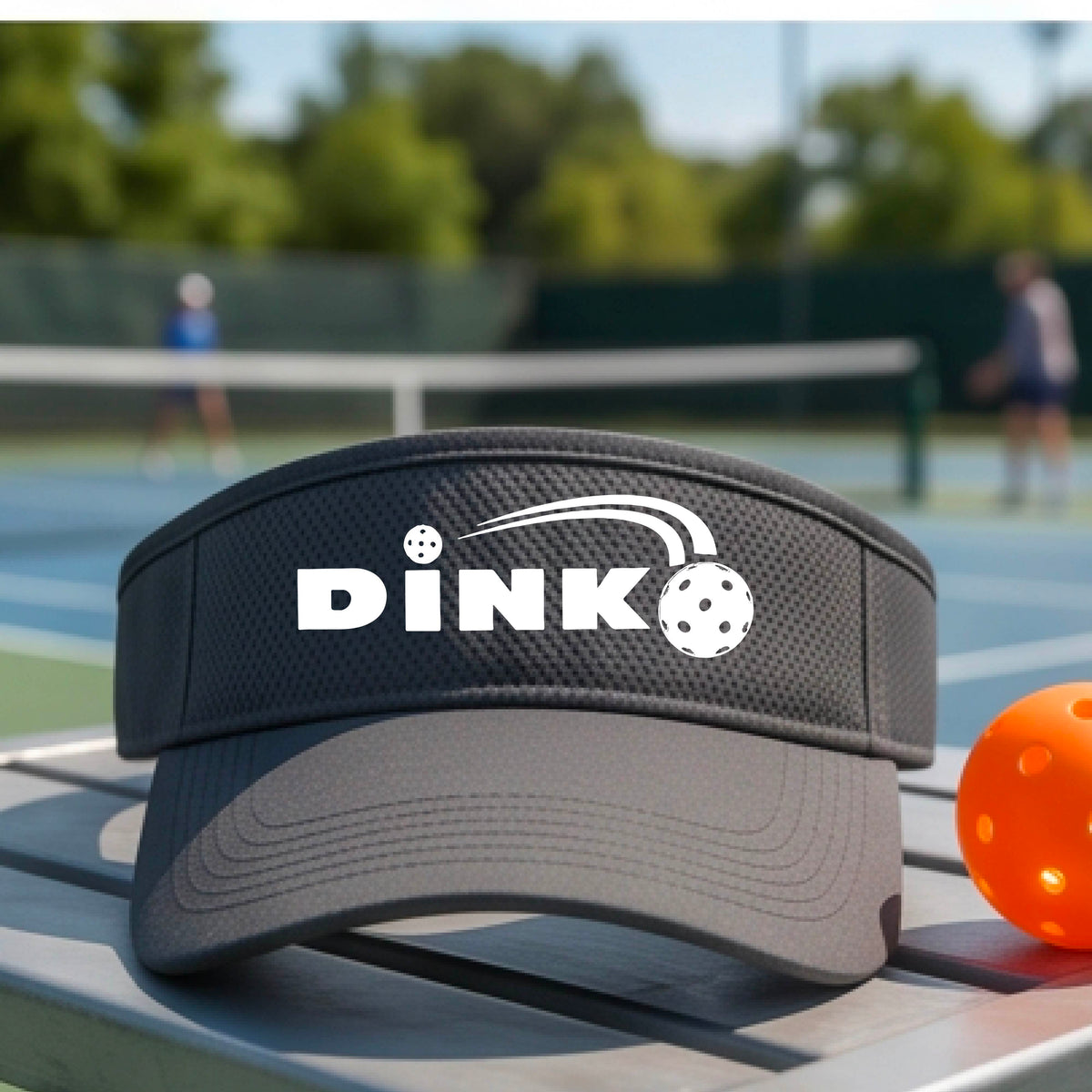 A close-up of a gray visor with a graphic of white 'DINK' and a dink pickleball logo printed in white on the front, set against a blurred outdoor pickleball court background.