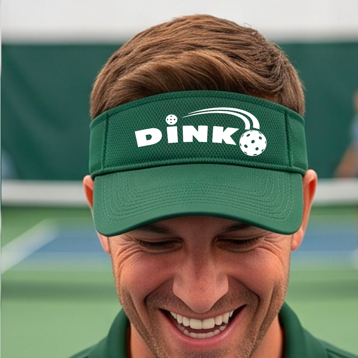 A close-up of a green visor with a graphic of white 'DINK' and a dink pickleball logo printed in white on the front, set against a blurred outdoor pickleball court background.