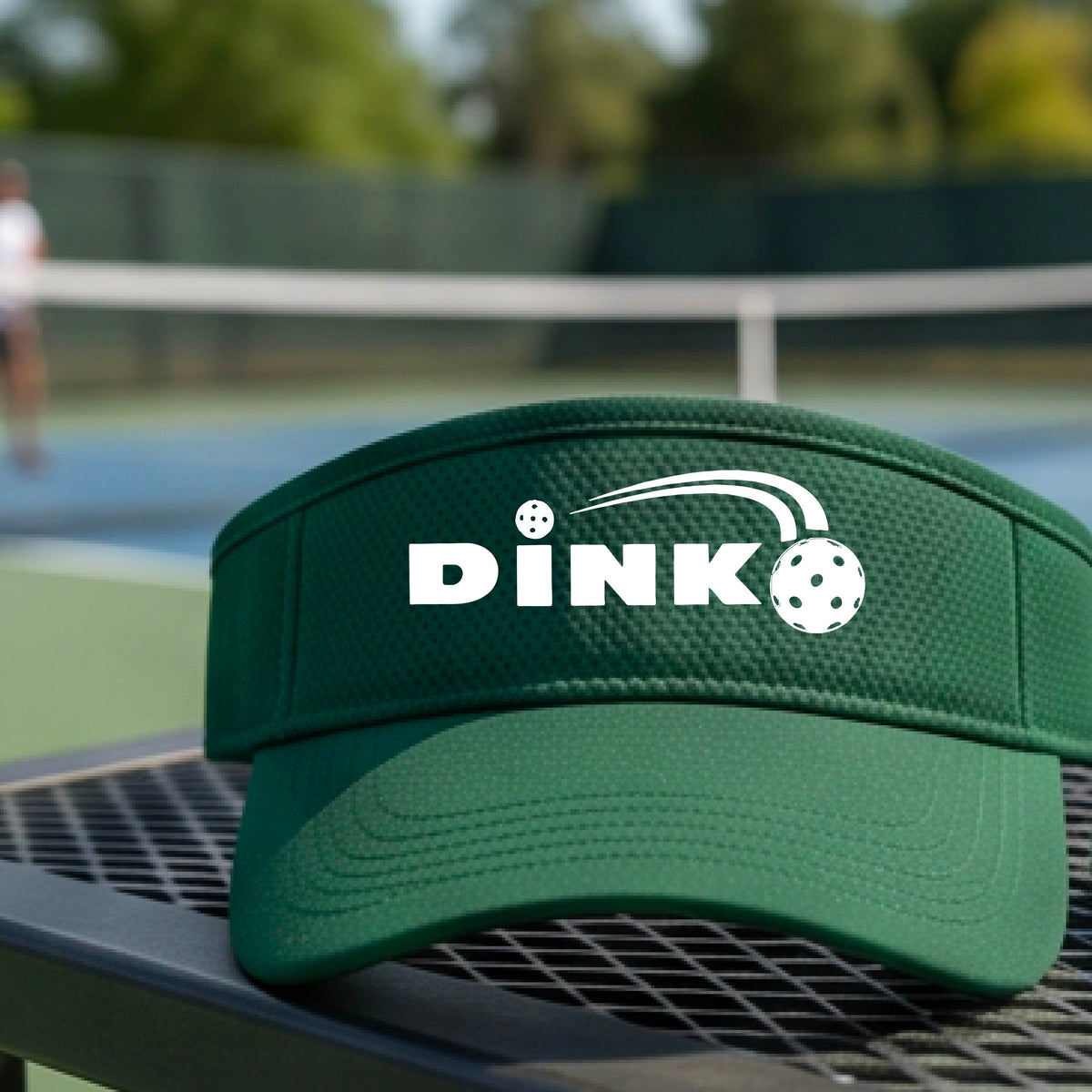 A close-up of a green visor with a graphic of white 'DINK' and a dink pickleball logo printed in white on the front, set against a blurred outdoor pickleball court background.