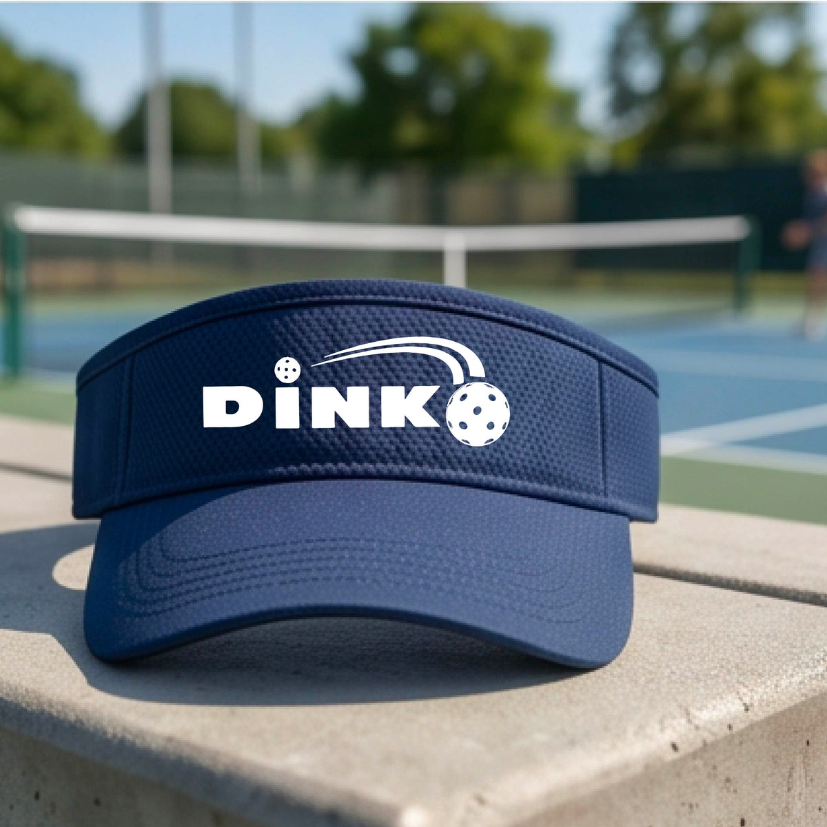 A close-up of a navy blue visor with a graphic of white 'DINK' and a dink pickleball logo printed in white on the front, set against a blurred outdoor pickleball court background.
