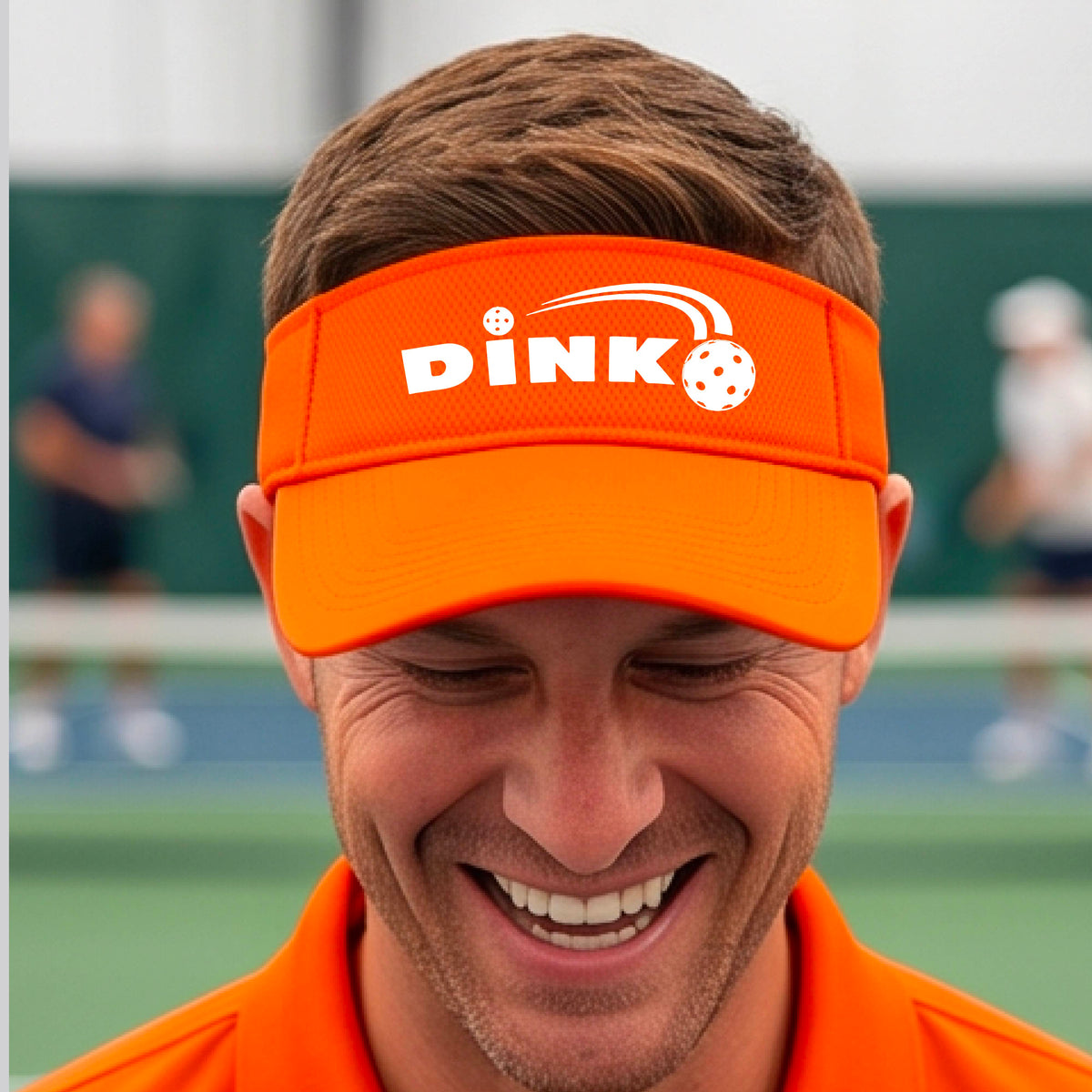 A close-up of an orange visor with a graphic of white 'DINK' and a dink pickleball logo printed in white on the front, set against a blurred outdoor pickleball court background.