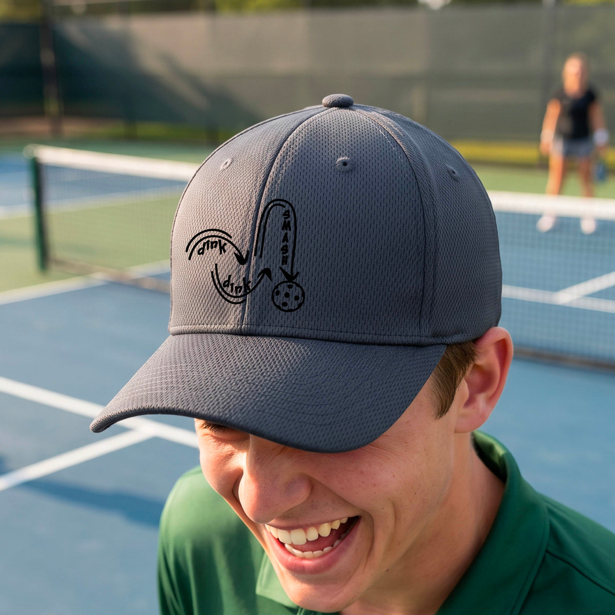 A close-up lifestyle shot of a gray athletic hat with a black pickleball 'DINK DINK SMASH' logo with a pickleball graphic