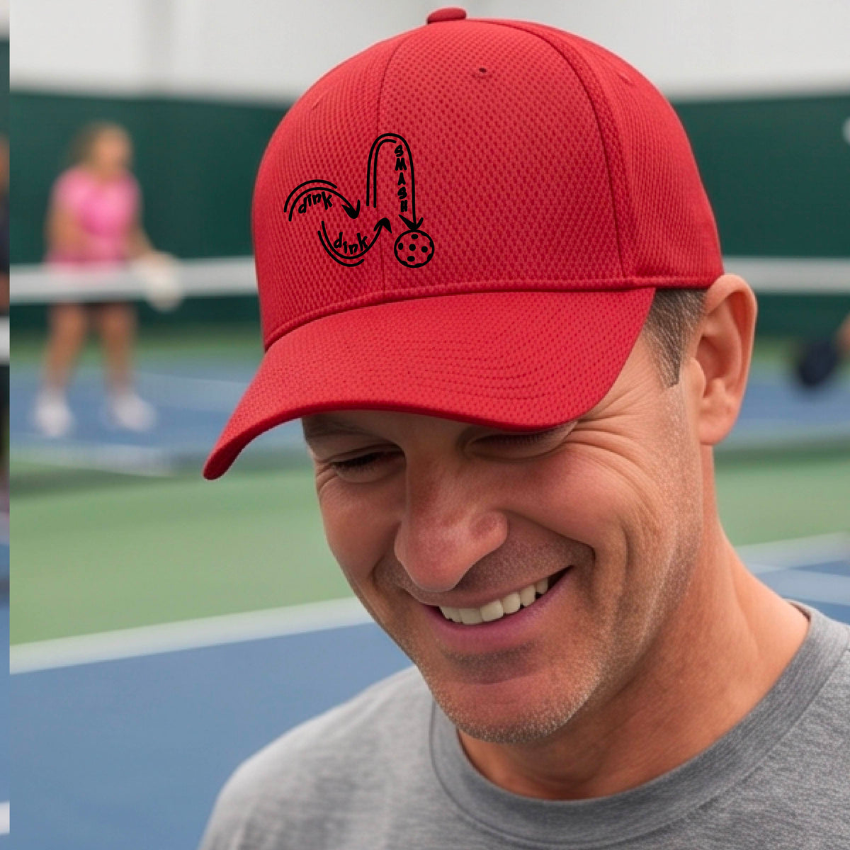 A close-up lifestyle shot of an red athletic hat with a black pickleball 'DINK DINK SMASH' logo with a pickleball graphic