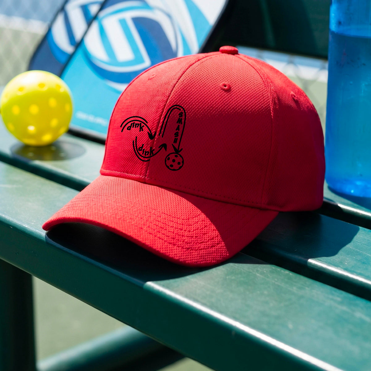 A close-up lifestyle shot of an red athletic hat with a black pickleball 'DINK DINK SMASH' logo with a pickleball graphic