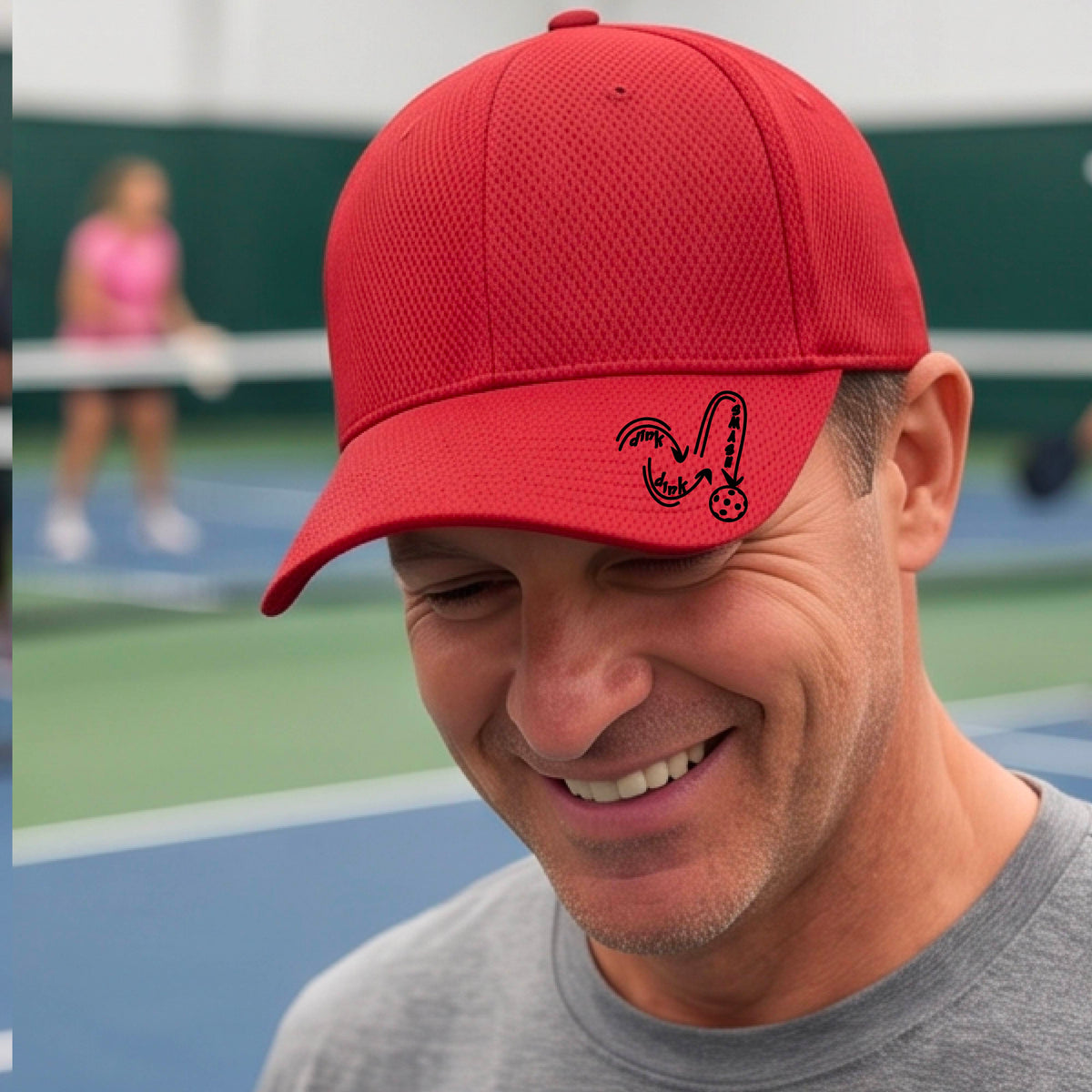 A close-up lifestyle shot of a red athletic hat with a black pickleball 'DINK DINK SMASH' logo with a pickleball graphic.
