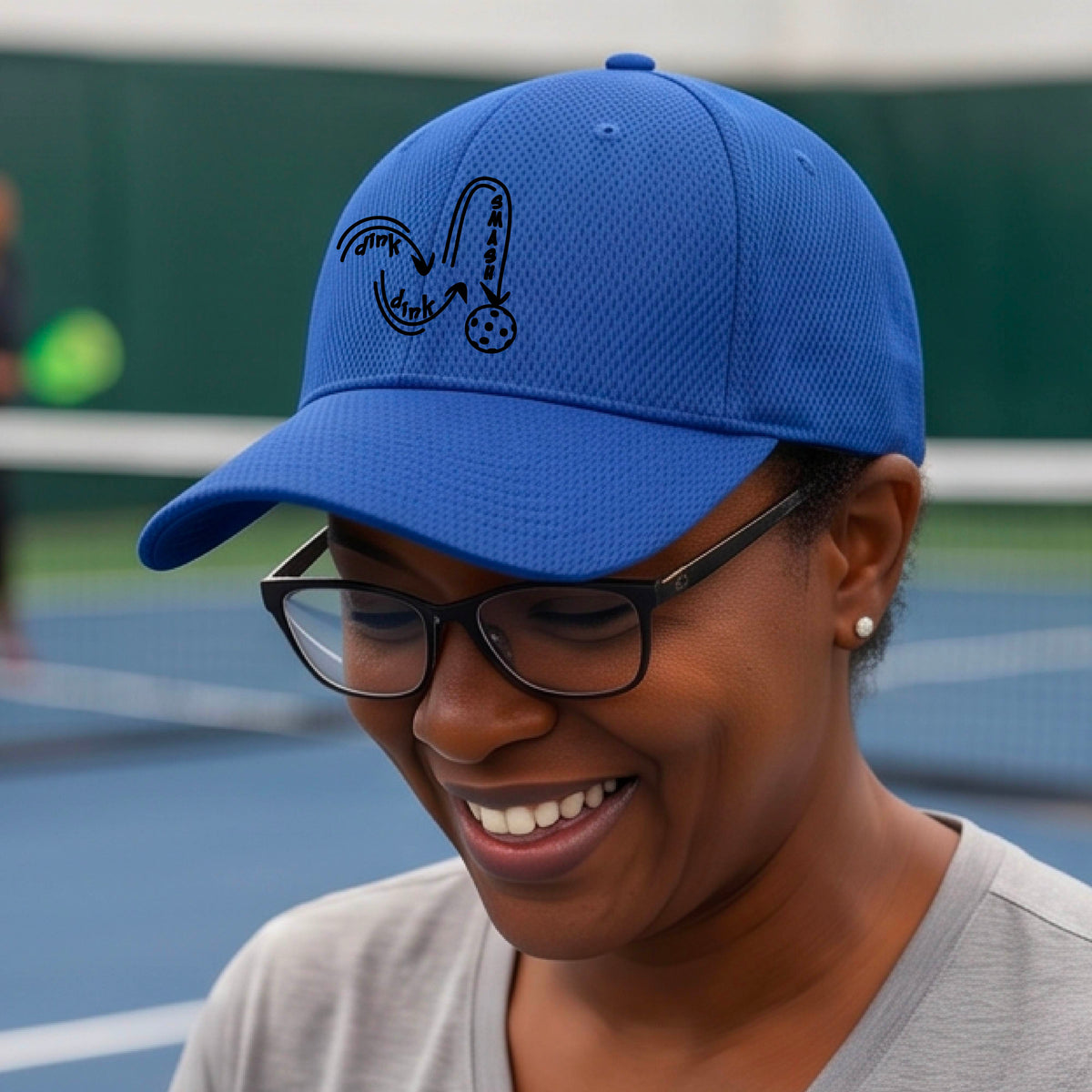 A close-up lifestyle shot of a royal blue athletic hat with a black pickleball 'DINK DINK SMASH' logo with a pickleball graphic.