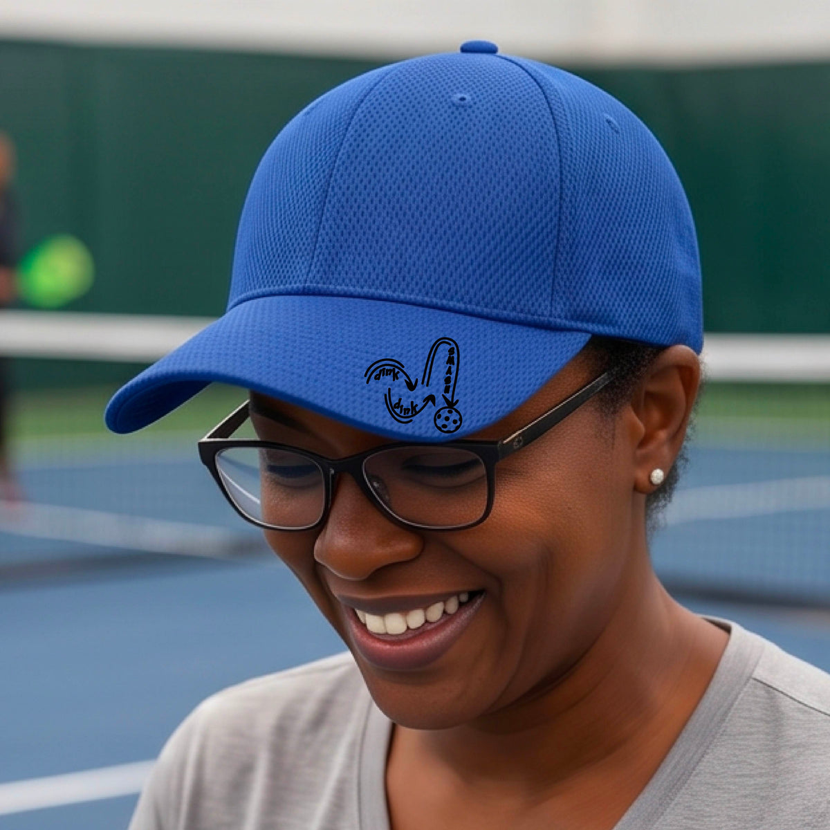 A close-up lifestyle shot of a royal blue athletic hat with a black pickleball 'DINK DINK SMASH' logo with a pickleball graphic.