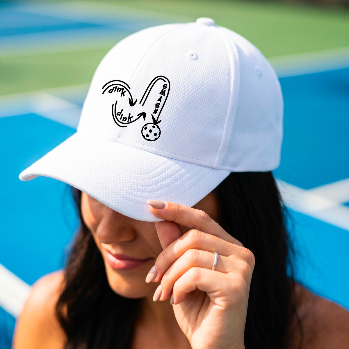 A close-up lifestyle shot of a White athletic hat with a black pickleball 'DINK DINK SMASH' logo with a pickleball graphic.