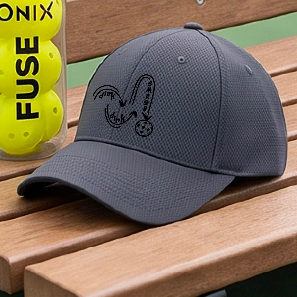 A close-up lifestyle shot of a gray athletic hat with a black pickleball 'DINK DINK SMASH' logo with a pickleball graphic