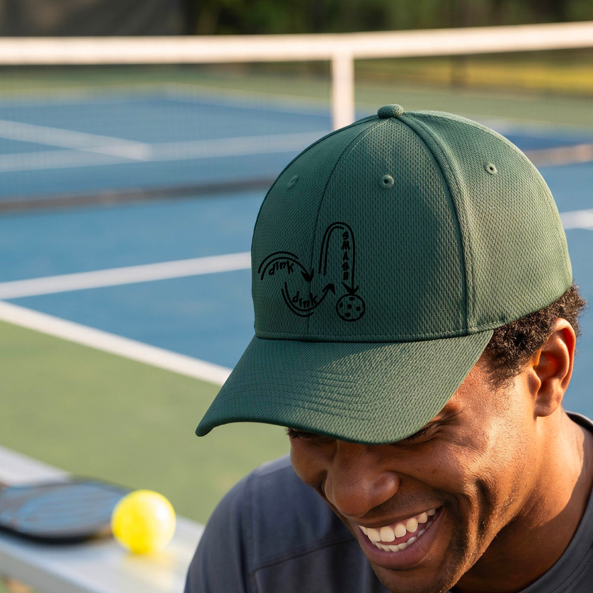 A close-up lifestyle shot of a green athletic hat with a black pickleball 'DINK DINK SMASH' logo with a pickleball graphic