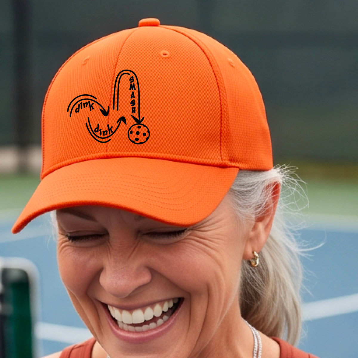 A close-up lifestyle shot of an orange athletic hat with a black pickleball 'DINK DINK SMASH' logo with a pickleball graphic