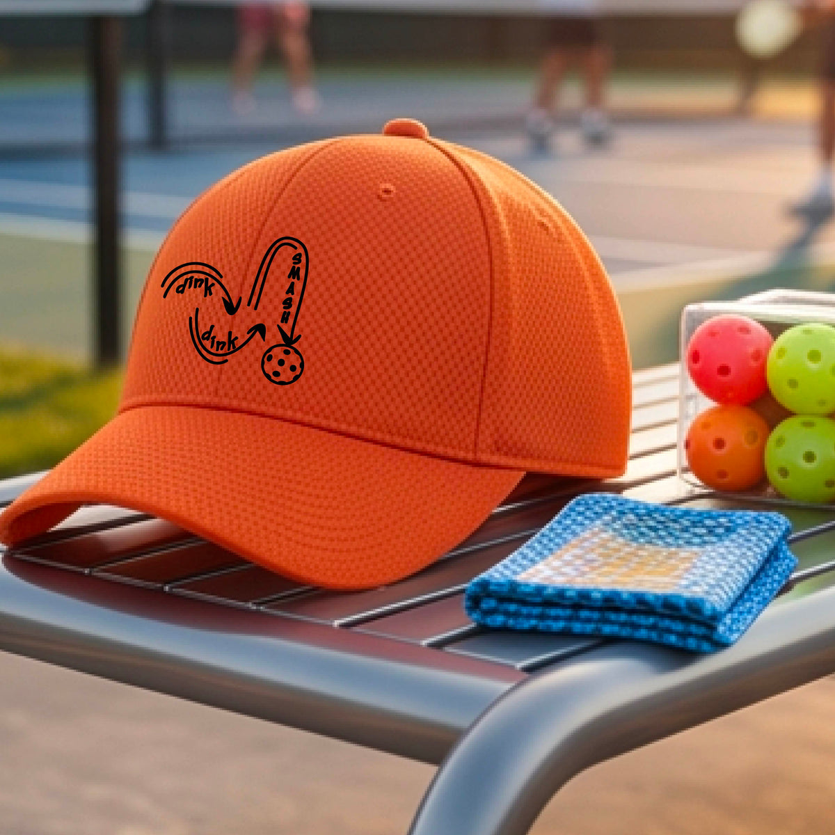 A close-up lifestyle shot of an orange athletic hat with a black pickleball 'DINK DINK SMASH' logo with a pickleball graphic