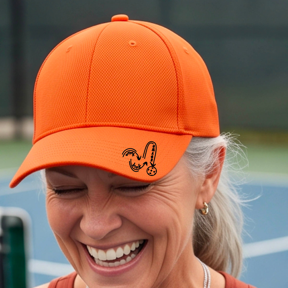 A close-up lifestyle shot of an orange athletic hat with a black pickleball 'DINK DINK SMASH' logo with a pickleball graphic