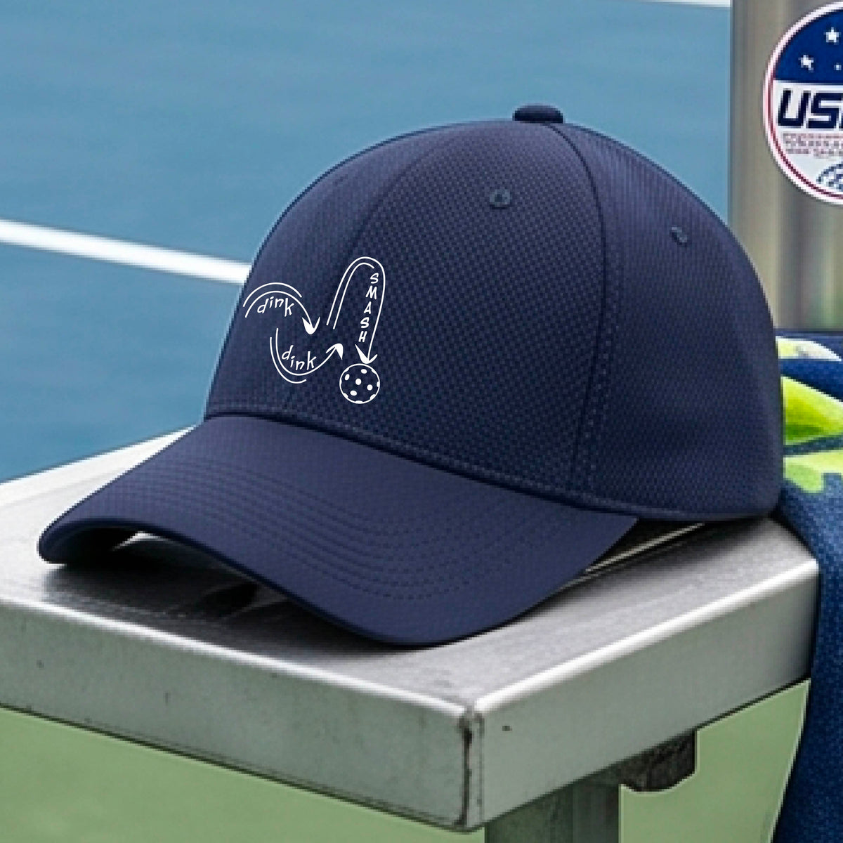 A close-up lifestyle shot of a navy blue athletic mid-profile hat with a white pickleball 'DINK DINK SMASH' logo with a pickleball graphic