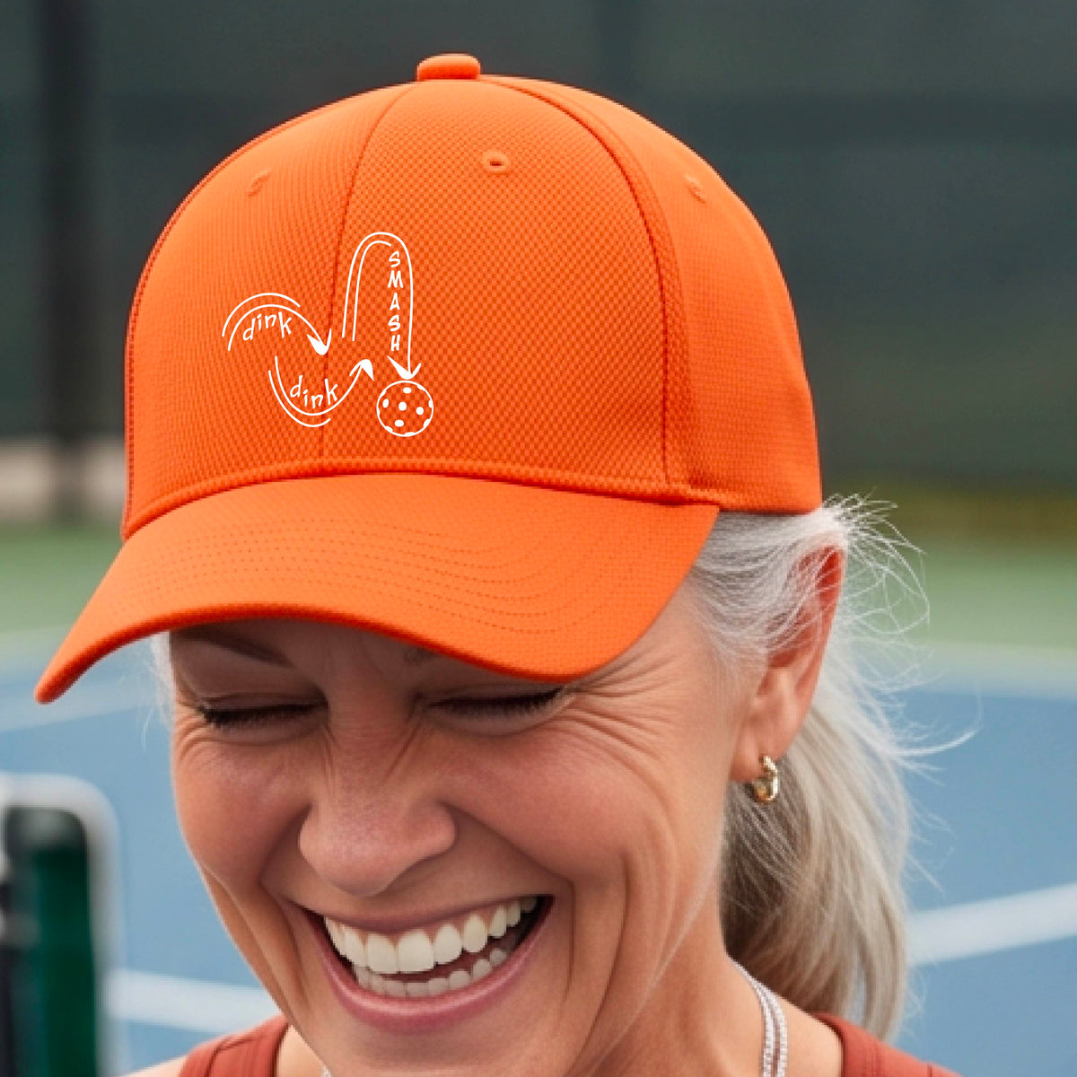 A close-up lifestyle shot of an orange athletic mid-profile hat with a white pickleball 'DINK DINK SMASH' logo with a pickleball graphic