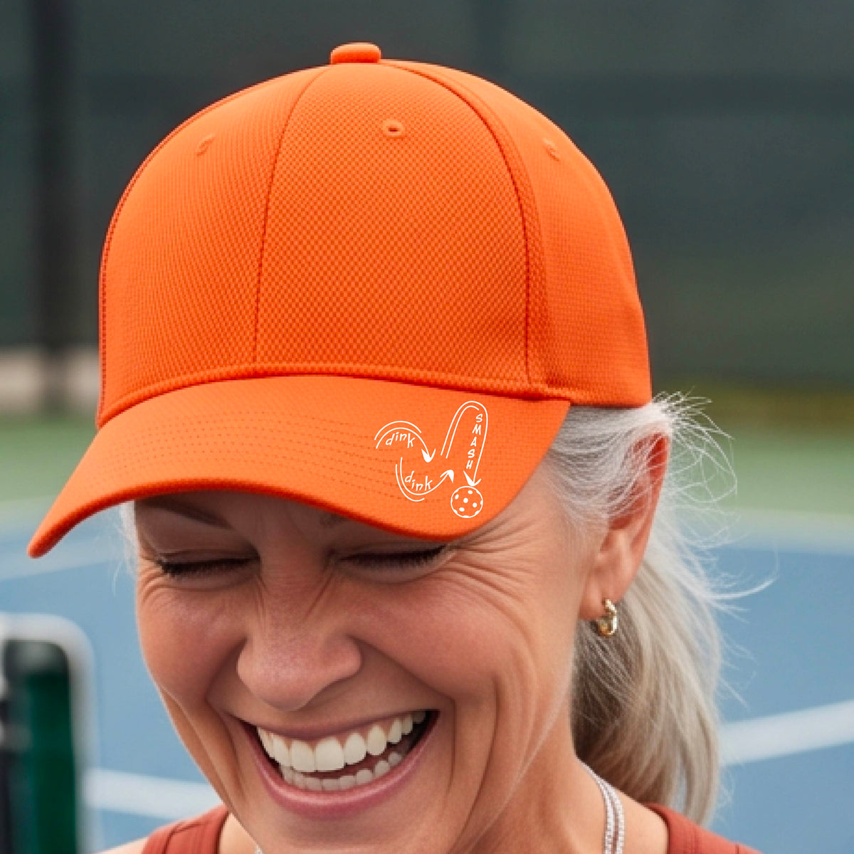 A close-up lifestyle shot of an orange athletic mid-profile hat with a white pickleball 'DINK DINK SMASH' logo with a pickleball graphic