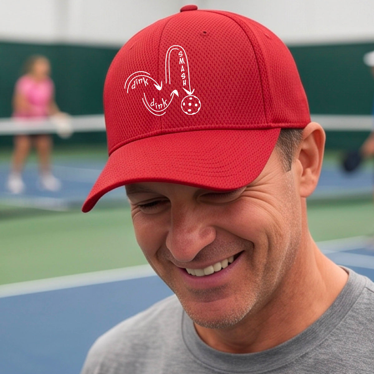 A close-up lifestyle shot of a red athletic mid-profile hat with a white pickleball 'DINK DINK SMASH' logo with a pickleball graphic
