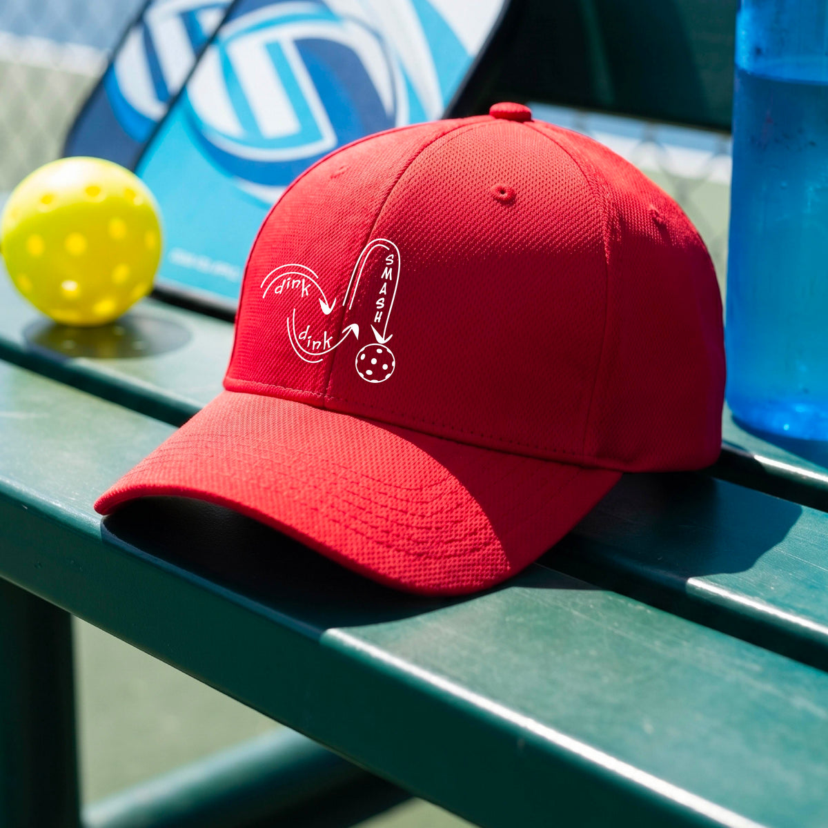 A close-up lifestyle shot of a red athletic mid-profile hat with a white pickleball 'DINK DINK SMASH' logo with a pickleball graphic