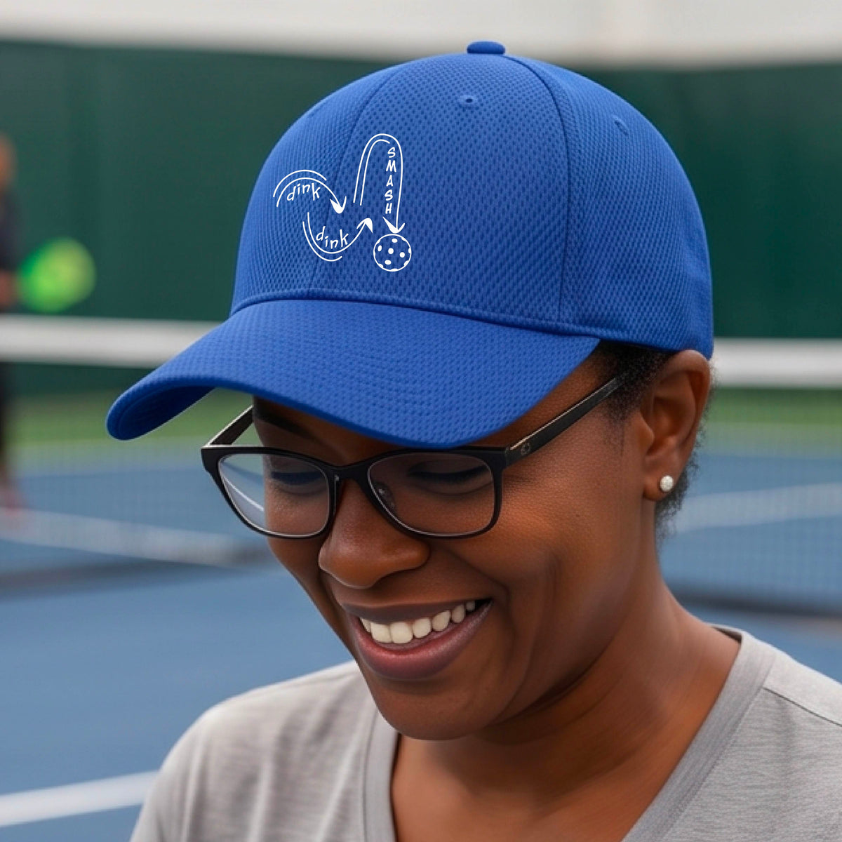 A close-up lifestyle shot of a royal blue athletic mid-profile hat with a white pickleball 'DINK DINK SMASH' logo with a pickleball graphic