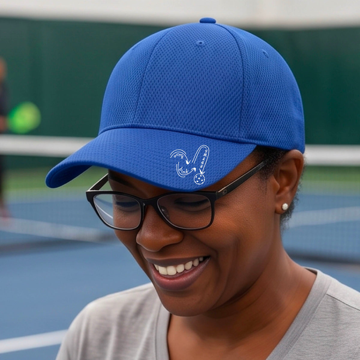A close-up lifestyle shot of a royal blue athletic mid-profile hat with a white pickleball 'DINK DINK SMASH' logo with a pickleball graphic