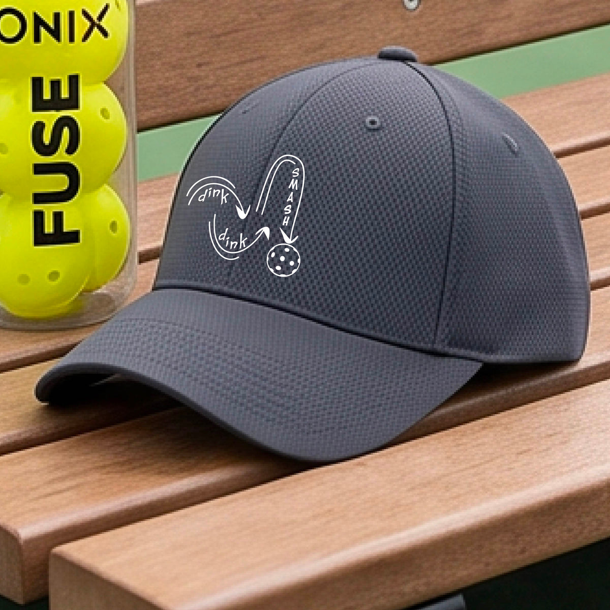 A close-up lifestyle shot of a gray athletic mid-profile hat with a white pickleball 'DINK DINK SMASH' logo with a pickleball graphic