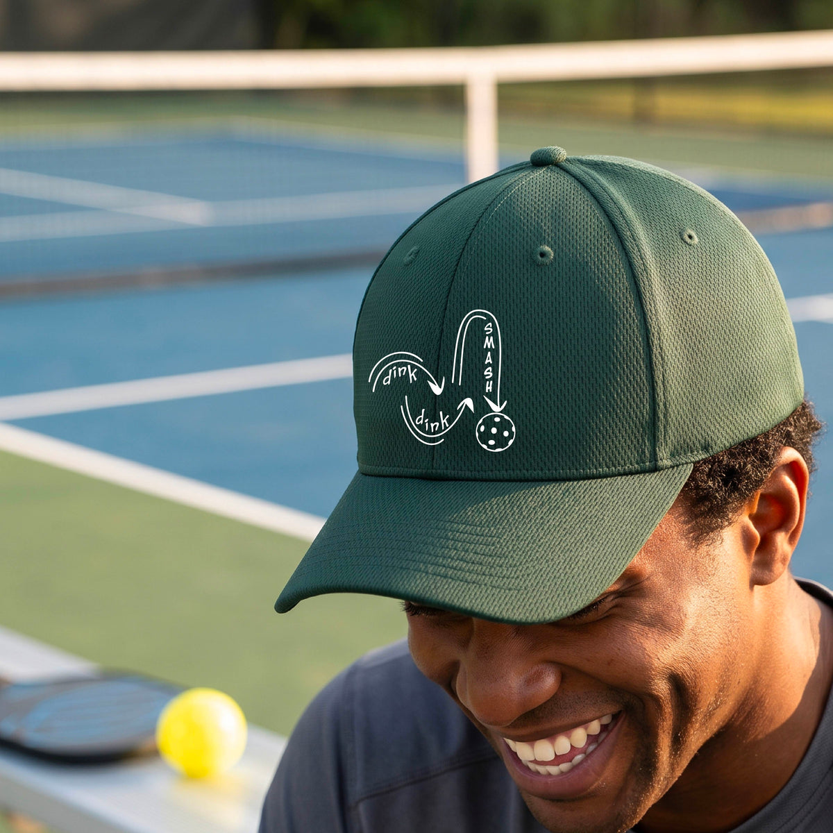 A close-up lifestyle shot of a green athletic mid-profile hat with a white pickleball 'DINK DINK SMASH' logo with a pickleball graphic