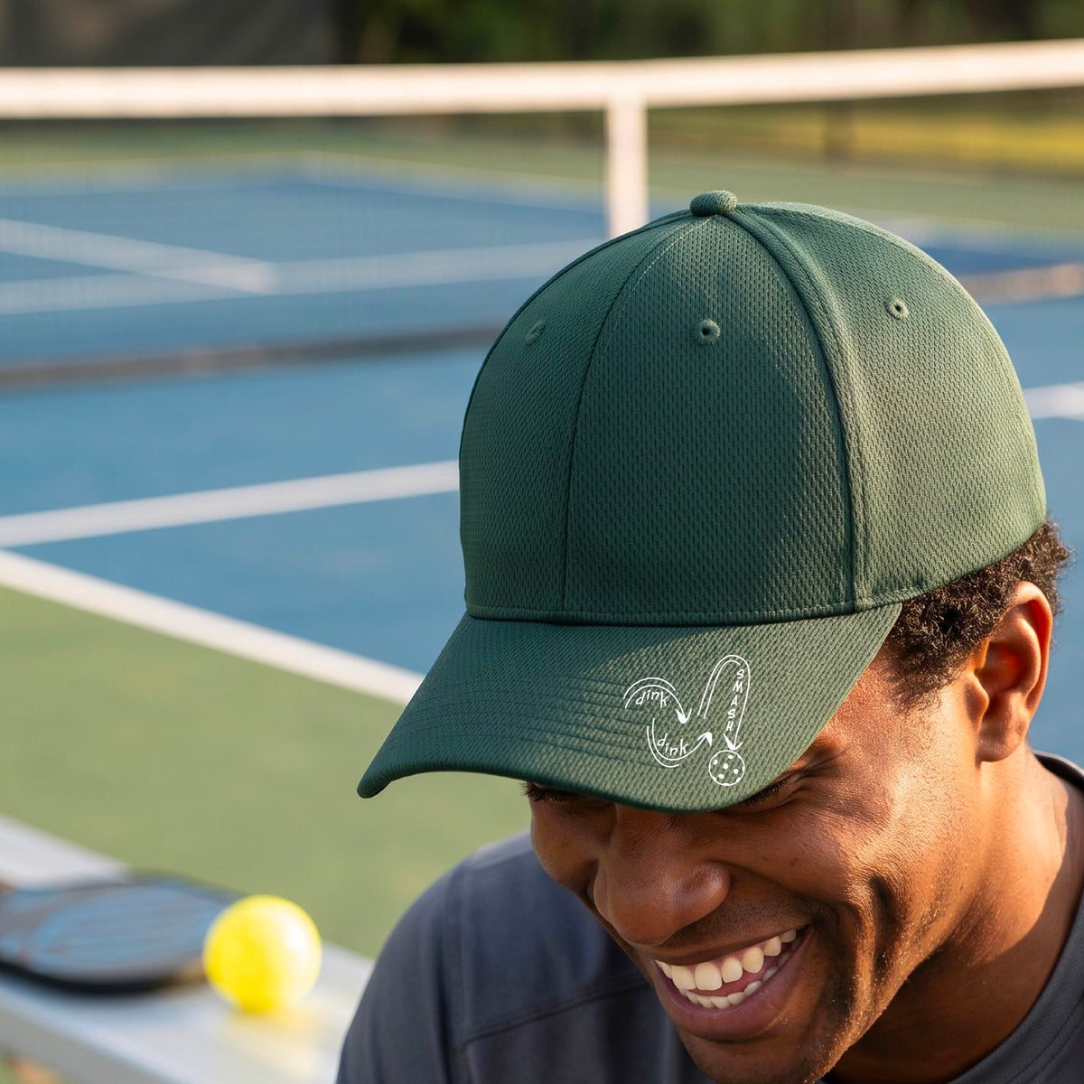 A close-up lifestyle shot of a green athletic mid-profile hat with a white pickleball 'DINK DINK SMASH' logo with a pickleball graphic