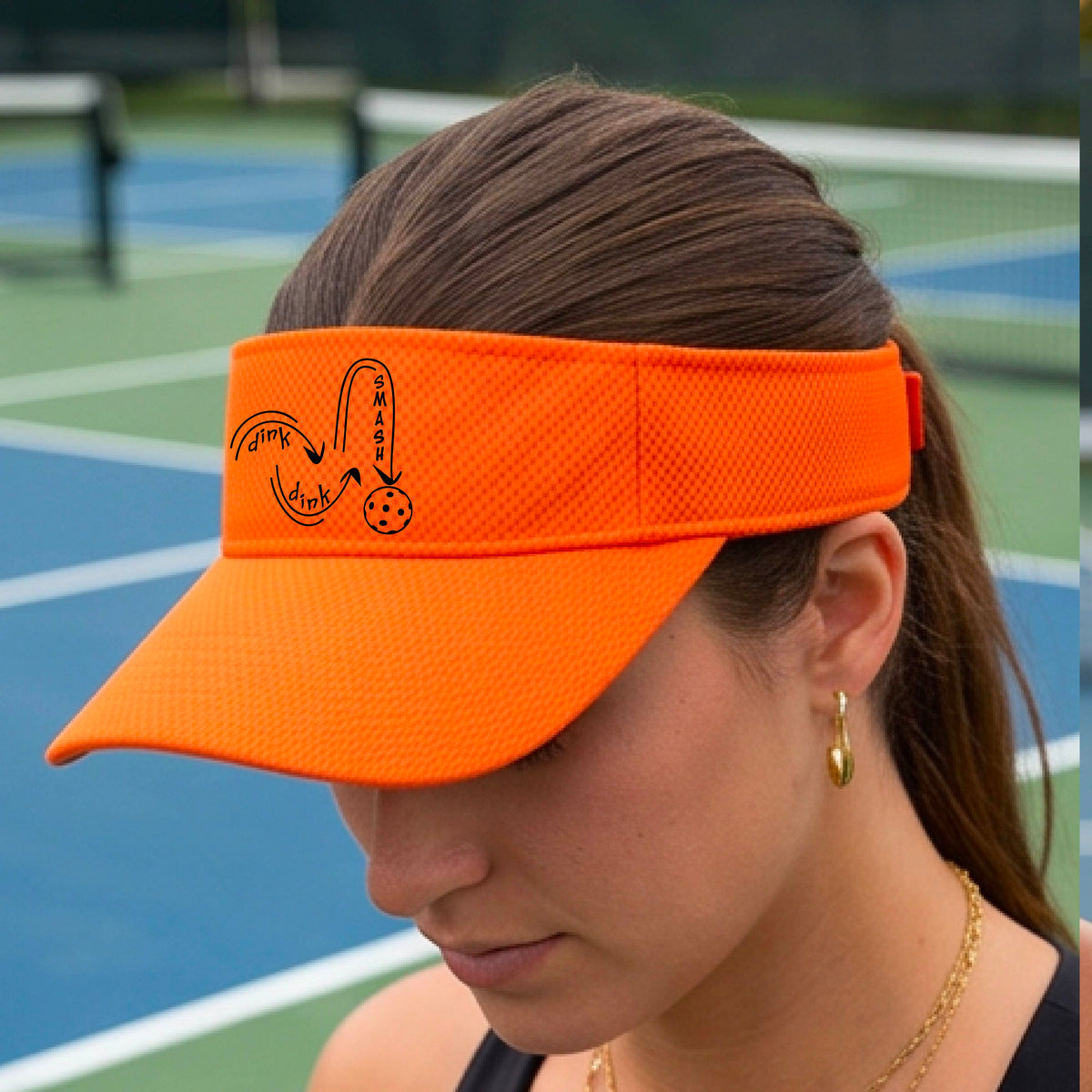 A close-up lifestyle shot of an orange athletic visor with a black pickleball 'DINK DINK SMASH' logo with a pickleball graphic.