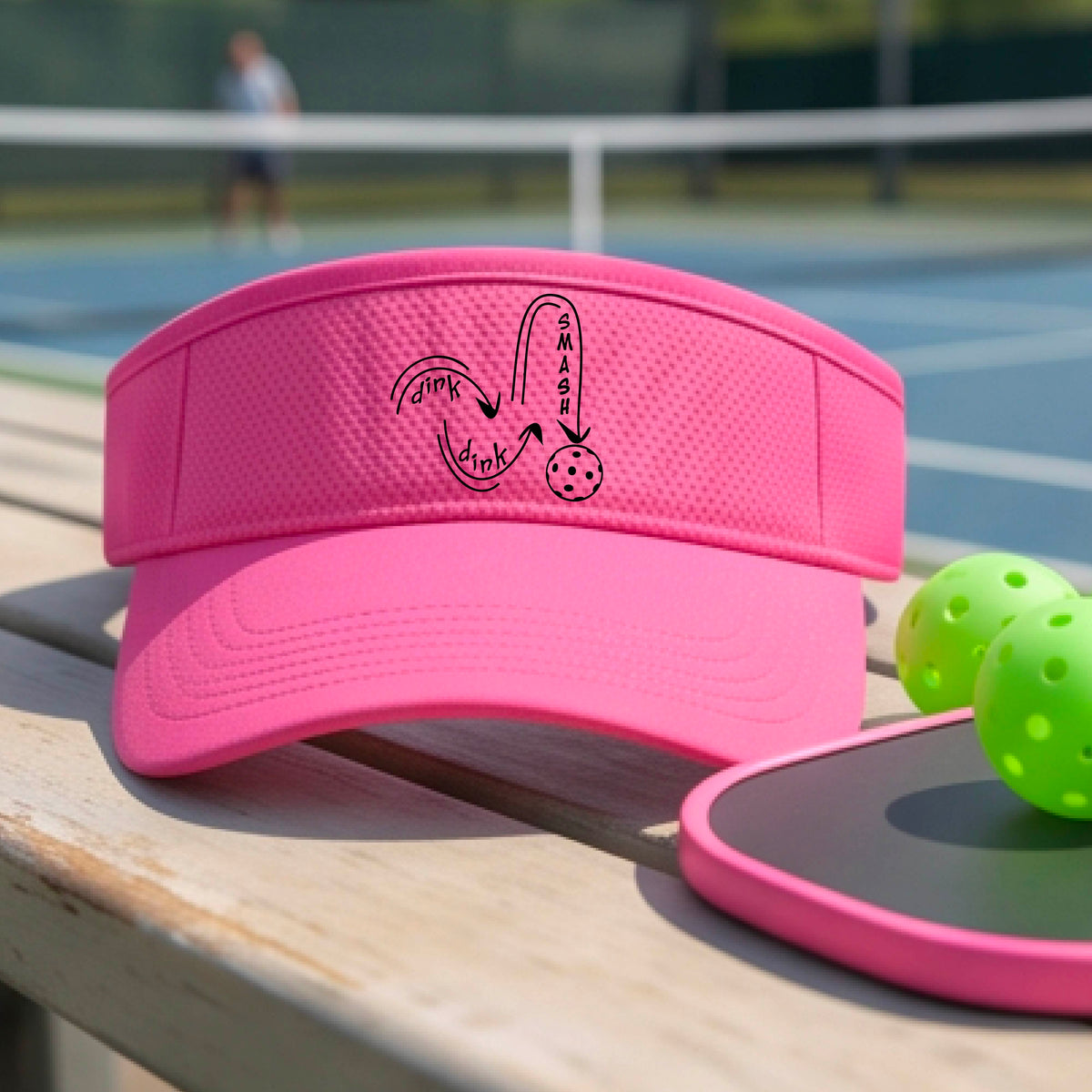 A close-up lifestyle shot of a pink athletic visor with a black pickleball 'DINK DINK SMASH' logo with a pickleball graphic.