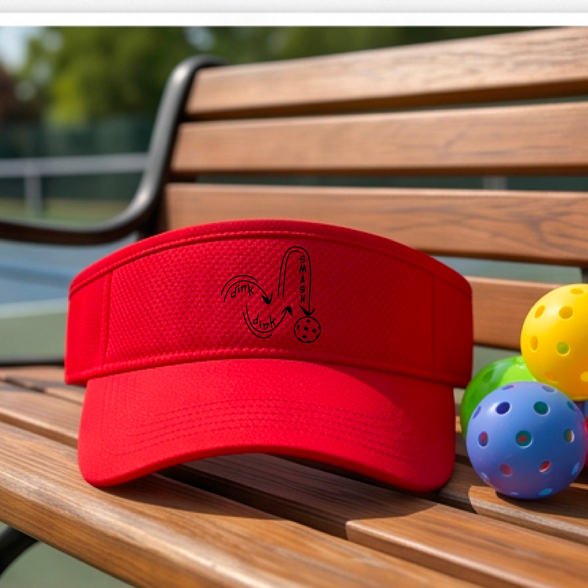 A close-up lifestyle shot of a red athletic visor with a black pickleball 'DINK DINK SMASH' logo with a pickleball graphic.