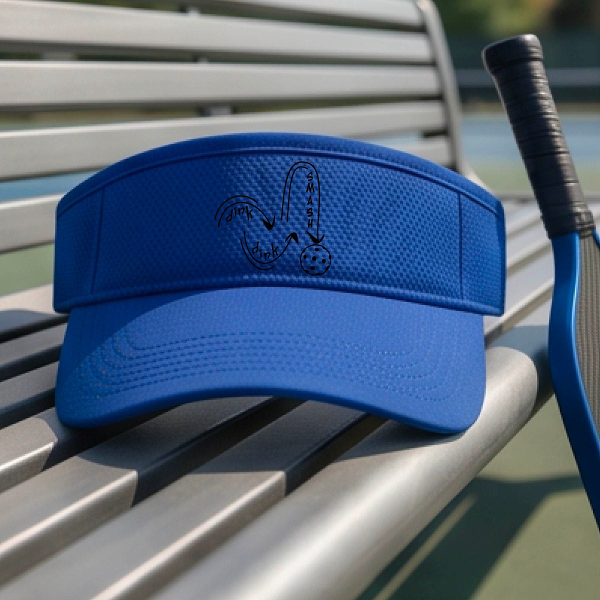 A close-up lifestyle shot of a royal blue athletic visor with a black pickleball 'DINK DINK SMASH' logo with a pickleball graphic.
