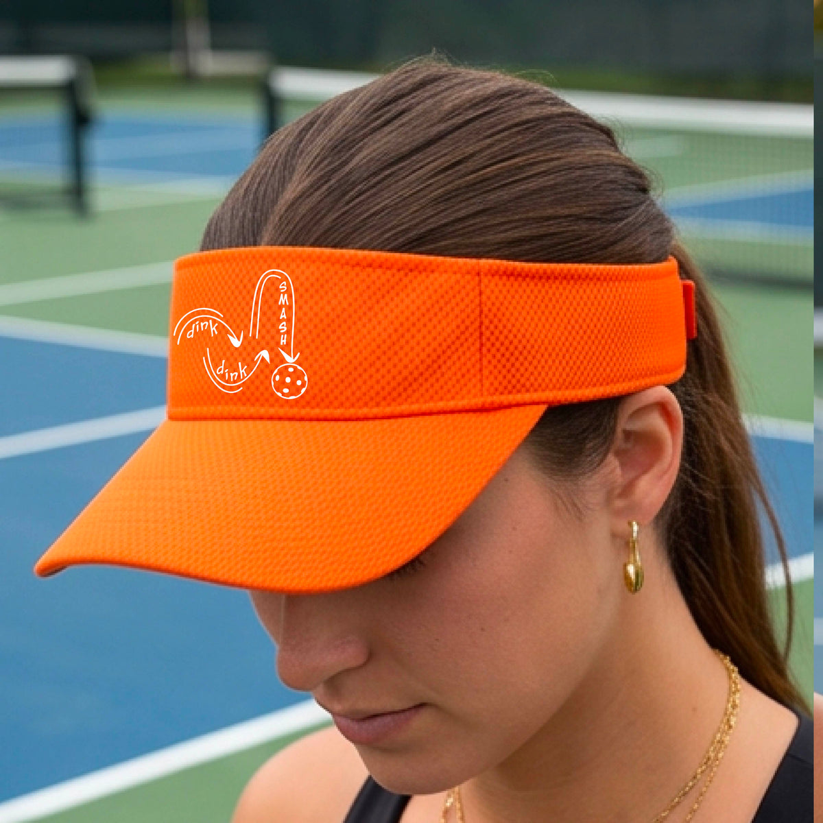 A close-up lifestyle shot of a bright orange athletic visor with a white pickleball 'DINK DINK SMASH' logo with a pickleball graphic