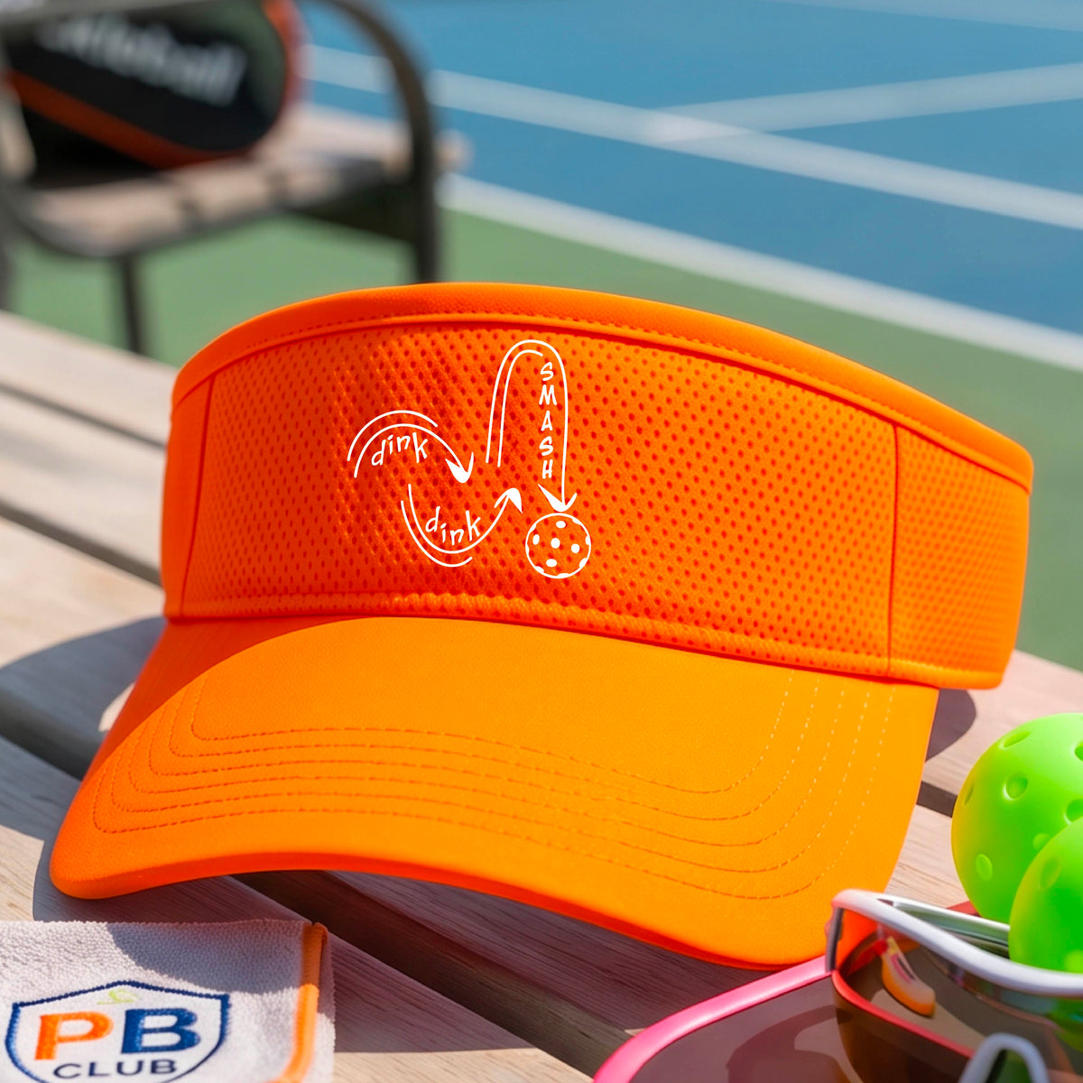A close-up lifestyle shot of a bright orange athletic visor with a white pickleball 'DINK DINK SMASH' logo with a pickleball graphic